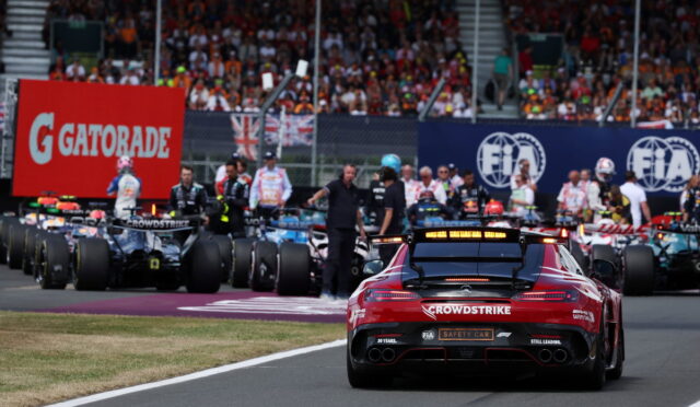 Rear view of a red safety car labeled 'CrowdStrike' on a race track, with race cars and a crowded grandstand in the background
