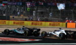 Two Formula 1 cars race side by side on a track, dust behind them, with bright Pirelli banners in the background and spectators beyond the fence.