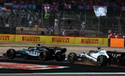 Two Formula 1 cars race side by side on a track, dust behind them, with bright Pirelli banners in the background and spectators beyond the fence.