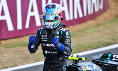 F1 driver in Petronas/Microsoft suit raises fists beside a Mercedes race car on track.
