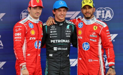 Three Formula 1 drivers pose together on a podium, left and right in red Ferrari suits and center in a black Mercedes Petronas suit, against a blue FIA backdrop.