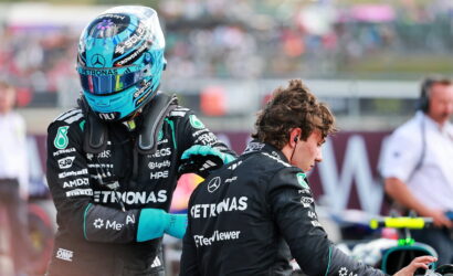 Two Mercedes Formula 1 team members in black Petronas suits, one wearing a blue helmet, stand in a busy pit area with a blurred crowd behind them.