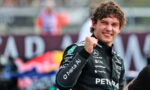 Smiling Formula 1 driver in a Petronas Mercedes racing suit raises a clenched fist in celebration on a blurred track backdrop.