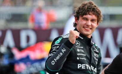 Smiling Formula 1 driver in a Petronas Mercedes racing suit raises a clenched fist in celebration on a blurred track backdrop.