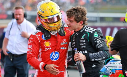 Two Formula 1 drivers in the pit lane chat, one in a red Ferrari suit with a yellow helmet and the other in a black Mercedes suit holding a water bottle.