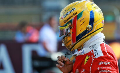 Side-profile of a race car driver wearing a yellow helmet and red racing suit with sponsor logos, adjusting his headset.