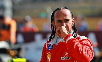 Male race car driver in red suit looks pensive, hand near mouth, paddock backdrop behind him.