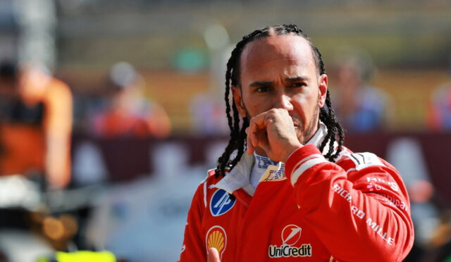 Male race car driver in red suit looks pensive, hand near mouth, paddock backdrop behind him.