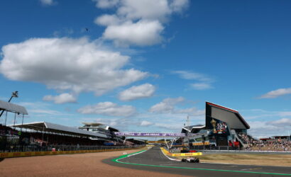 Race car on a curving asphalt track at a large grand prix stadium under a blue sky with white clouds.