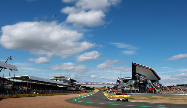 Race car on a curving asphalt track at a large grand prix stadium under a blue sky with white clouds.