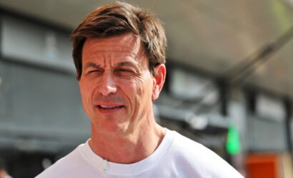 Middle-aged man with short brown hair squinting in bright sunlight, wearing a white crew-neck shirt outdoors near a building.