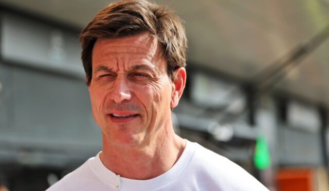 Man in a white shirt outdoors squinting in bright light, with a worried expression and wind-swept hair.