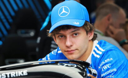 Young man in a blue Mercedes cap and team jersey leaning on a black race car, looking at the camera.