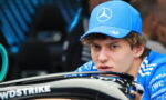 Young race car driver in a blue Mercedes cap and shirt looking over a race car cockpit with sponsor logos visible.