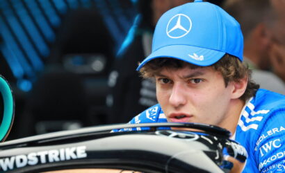 Young race car driver in a blue Mercedes cap and shirt looking over a race car cockpit with sponsor logos visible.