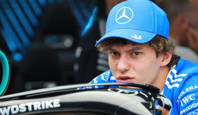 Young race car driver in a blue Mercedes cap and shirt looking over a race car cockpit with sponsor logos visible.