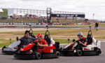Three go-karts racing side by side on a track, drivers smiling with a crowded pit area and TAG Heuer banners in the background.