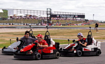 Three go-karts racing side by side on a track, drivers smiling with a crowded pit area and TAG Heuer banners in the background.