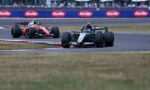Two Formula 1 cars race around a track corner, with a black front car and a red car close behind; Barilla banners line the infield.