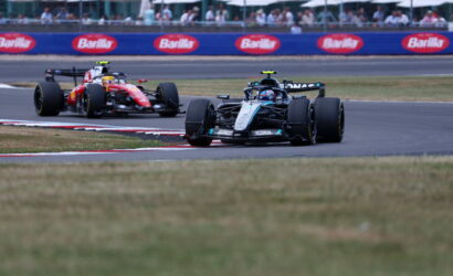 Two Formula 1 cars race around a track corner, with a black front car and a red car close behind; Barilla banners line the infield.