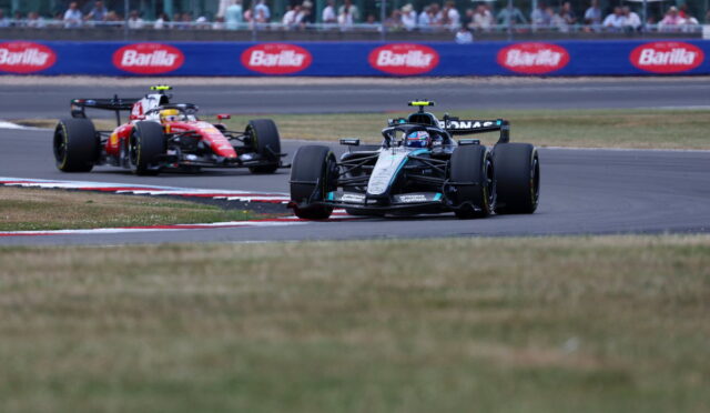 Two Formula 1 cars race around a track corner, with a black front car and a red car close behind; Barilla banners line the infield.
