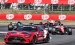Red Mercedes AMG safety car leading a Formula 1 race on a curving track, with bright red KitKat banners in the background and pursuing F1 cars behind.