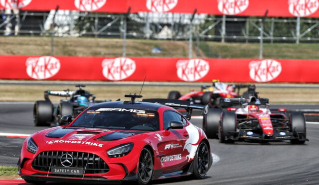 Red Mercedes AMG safety car leading a Formula 1 race on a curving track, with bright red KitKat banners in the background and pursuing F1 cars behind.