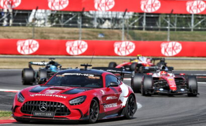 Red Mercedes safety car leads a pack of Formula 1 cars on a curving race track, with bright sponsor banners in the background.
