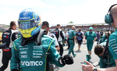 Formula 1 driver in teal Aramco racing suit and blue-yellow helmet gives a thumbs-up to a crew member in a crowded pit area.