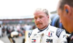 Male race car driver with blond hair and mustache in a white racing suit, standing in the pit area and looking thoughtful.