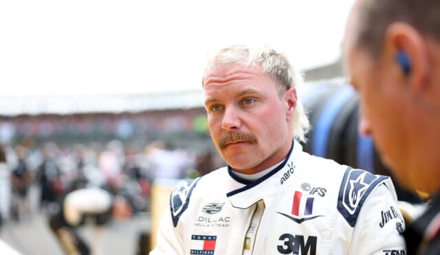 Male race car driver with blond hair and mustache in a white racing suit, standing in the pit area and looking thoughtful.