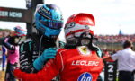 Two Formula 1 drivers in helmets share a hug on the track, one in blue Petronas gear and the other in red Leclerc Ferrari gear, with spectators in the background.