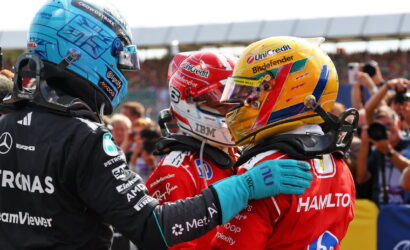 Three Formula 1 drivers in colorful helmets greet and embrace each other on the track as fans photograph in the background.