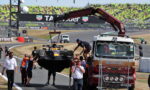 Race car being lifted from a tow truck by a red crane as crew stand by at a race track, with grandstands full of spectators in the background.