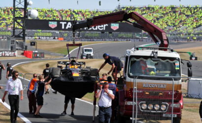 Race car being lifted from a tow truck by a red crane as crew stand by at a race track, with grandstands full of spectators in the background.
