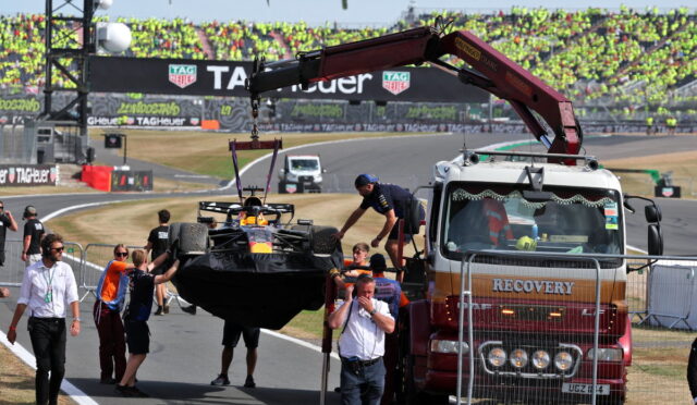 Race car being lifted from a tow truck by a red crane as crew stand by at a race track, with grandstands full of spectators in the background.