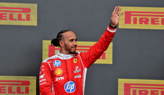 Ferrari driver in a red race suit waves from a podium with Pirelli logos in the background.