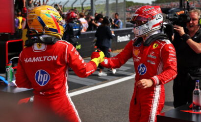 Two Formula 1 drivers in red Ferrari racing suits exchange a fist bump on the pit wall, with sponsor logos visible and a crowd in the background.