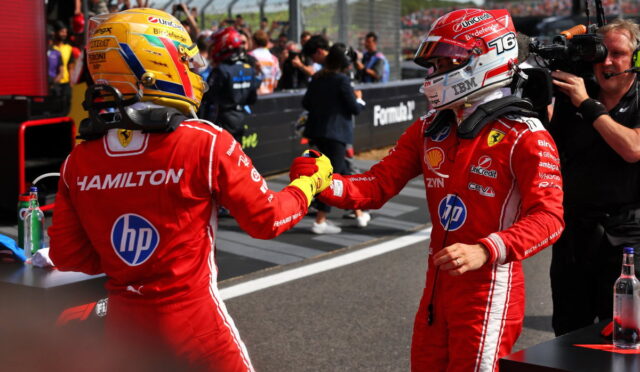 Two Formula 1 drivers in red Ferrari racing suits exchange a fist bump on the pit wall, with sponsor logos visible and a crowd in the background.