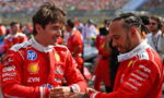 Two Ferrari drivers in red racing suits greet each other with a handshake, smiling, on a busy race track backdrop.
