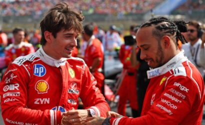 Two Ferrari drivers in red racing suits greet each other with a handshake, smiling, on a busy race track backdrop.