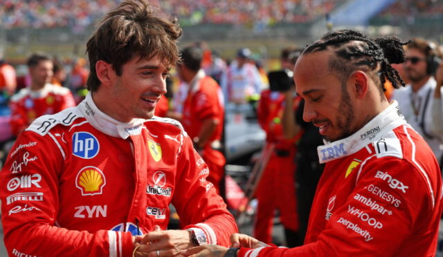 Two Ferrari drivers in red racing suits greet each other with a handshake, smiling, on a busy race track backdrop.