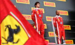 Two Formula 1 drivers in red Ferrari-style racing suits stand on a podium with Pirelli banners in the background and a Ferrari flag in the foreground.