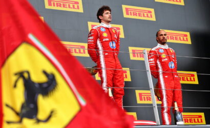 Two Formula 1 drivers in red Ferrari-style racing suits stand on a podium with Pirelli banners in the background and a Ferrari flag in the foreground.