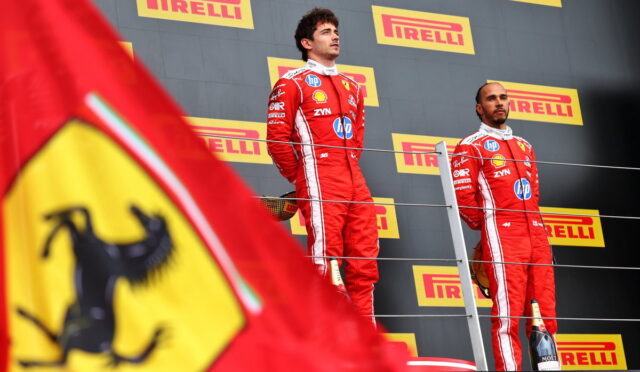 Two Formula 1 drivers in red Ferrari-style racing suits stand on a podium with Pirelli banners in the background and a Ferrari flag in the foreground.