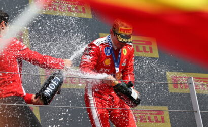 Ferrari driver on a podium spraying champagne, teammate holding a Moët bottle; sponsor banners in the background.