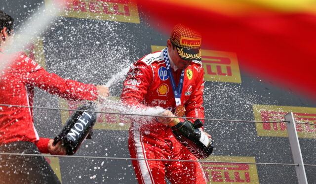Ferrari driver on a podium spraying champagne, teammate holding a Moët bottle; sponsor banners in the background.