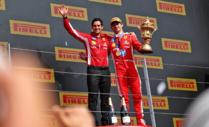 Two racecar drivers in red suits stand on a podium; one holds a large trophy and medals, with Pirelli banners behind them and champagne bottles at their feet.