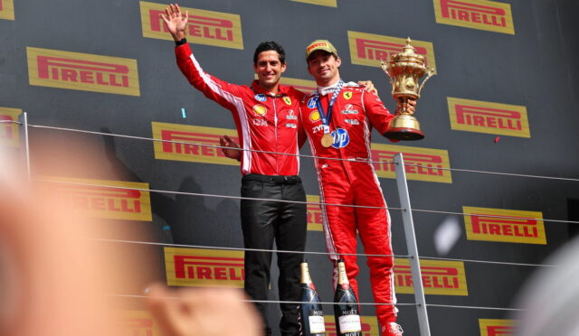 Two racecar drivers in red suits stand on a podium; one holds a large trophy and medals, with Pirelli banners behind them and champagne bottles at their feet.