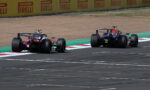 Two Formula 1 cars race side by side on a track, with yellow Pirelli barriers in the background.
